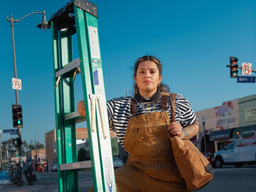 Johanna Toruño stands outside on a sidewalk. She is holding an upright ladder with her right hand; she carries a large tote bag on her left arm.