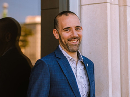 A white man wearing a blazer and button-up shirt is smiling while standing in front of a window.