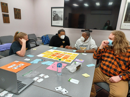 Four college students sitting around a table in a classroom with board game pieces on the table.