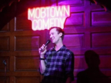 Dan stands smiling wearing a plaid shirt and holding a microphone in front of a sign that reads "mobtown comedy".