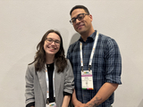 Two smiling humans one with glasses and shoulder length dark hair wearing a black shirt and grey blazer, and one with glasses and short dark hair wearing a lanyard and blue plaid shirt