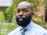 A person with a shaved head and beard wearing a blue shirt and a green and blue plaid tie.