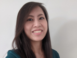 Portrait of Asian American woman with long dark brown hair in a green dress shirt smiling at the camera with a light beige wall in the background
