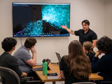 Student presents at a lab meeting. The screen shows an image from OpenSpace, a program that visualizes the universe in 3D.