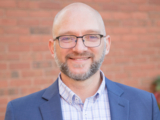 headshot of man wearing glasses, button down shirt, and blue blazer.