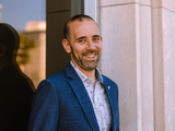 smiling man with close cropped hair and beard standing outside a building, in front of a window