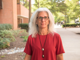 A portrait of Professor Denise Meringolo, wearing a red top and glasses, in an outdoor setting with a building, a pathway, and greenery in the background