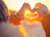 hands in mittens forming heart shape with sun setting in background